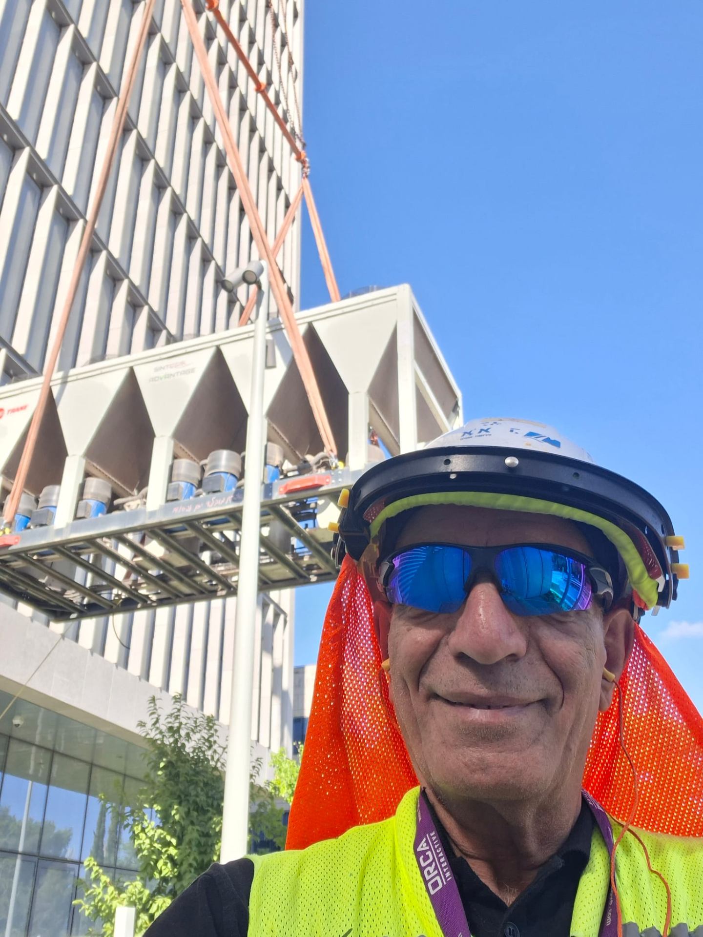 חנניה כהן בקסדה ובאפוד זוהר באתר בנייה על רקע מנוף ומזגן מורם